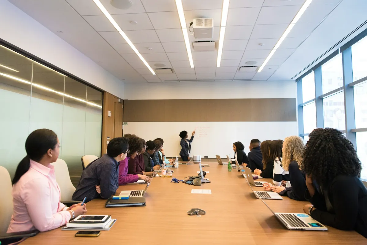 A group of colleagues around a boardroom table in discussion, one of them presenting at a whiteboard.